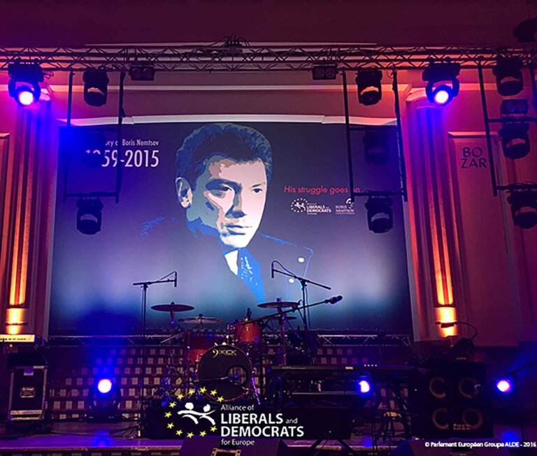 Backdrop en tissu imprimé pour scène de concert, affichant un portrait stylisé d’un homme avec la mention '1959–2015' et 'His struggle goes on', installé dans un lieu de spectacle éclairé par des projecteurs.