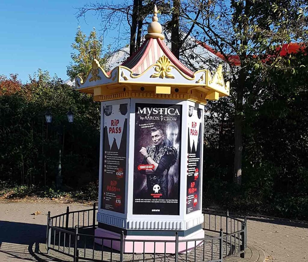 Affiches imprimées sur vinyle auto-adhésif collées sur un kiosque de parc d'attractions, présentant une promotion pour 'Mystica by Aaron Crow'