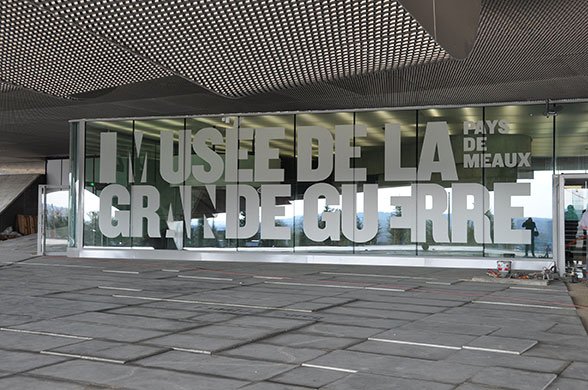 Entrée du Musée de la Grande Guerre à Meaux, avec une façade vitrée recouverte de vinyle sablée affichant le nom du musée en grandes lettres. L'aspect mat des lettres permet une lecture claire tout en filtrant la lumière. Le toit en structure métallique ajourée et le sol en dalles grises complètent l’esthétique contemporaine du bâtiment.