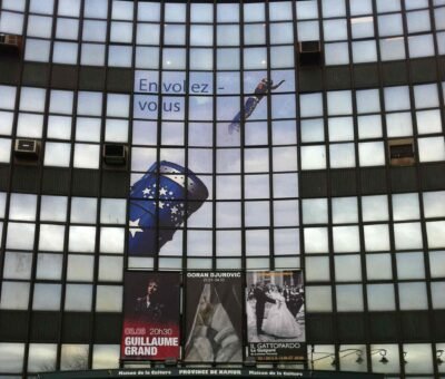 Impression sur vinyle pour façades : affichages publicitaires et événements culturels Façade en verre d’un bâtiment culturel ornée d’une grande impression vinyle représentant un sauteur en parachute en vol, accompagné du slogan « En volez-vous ». Un chapeau bleu étoilé est également visible, symbolisant le thème de l’événement. Des affiches de spectacle sont accrochées en dessous.