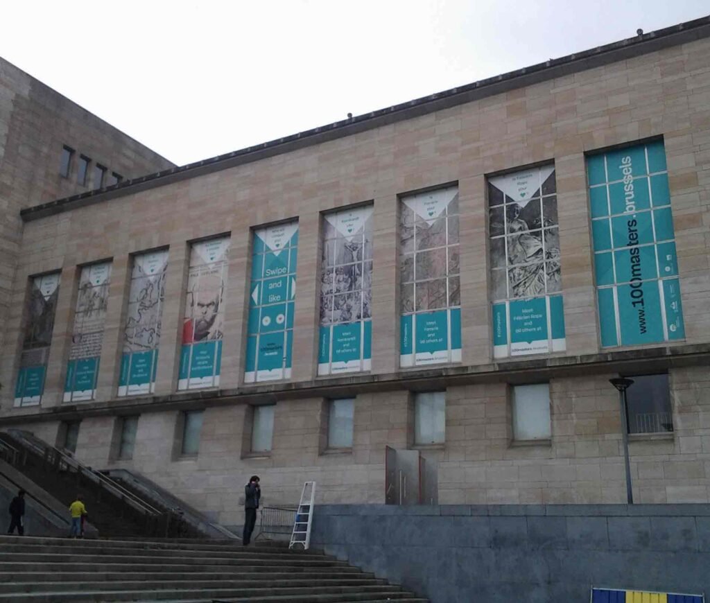 Façade du bâtiment de la Bibliothèque Royale de Bruxelles, décorée de grandes impressions vinyle sur les fenêtres. Les bannières montrent des illustrations artistiques, des portraits historiques et des messages interactifs comme « Swipe and like », promouvant l’exposition « 100masters.brussels ».