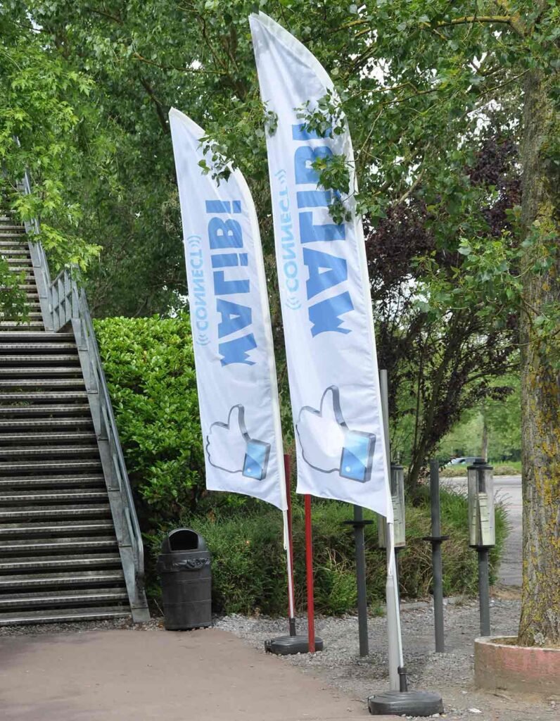 Deux drapeaux publicitaires en tissu léger, blancs avec texte en cyrillique et icône de "like", plantés sur des poteaux dans un espace extérieur verdoyant près d’un escalier métallique. Beach flag.