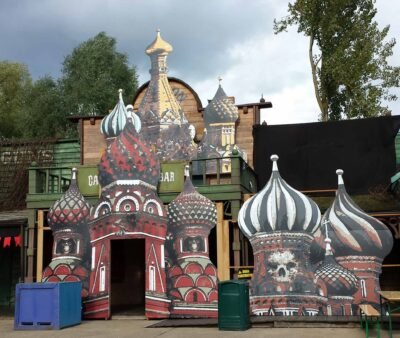 écor en forex Halloween – Dômes fantomatiques inspirés de la cathédrale Saint-Basile Décoration d’extérieur en forex pour Halloween représentant des dômes russes stylisés avec motifs macabres et crânes, installée devant un bar dans un parc festif.