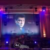 Backdrop en tissu imprimé pour scène de concert, affichant un portrait stylisé d’un homme avec la mention '1959–2015' et 'His struggle goes on', installé dans un lieu de spectacle éclairé par des projecteurs.