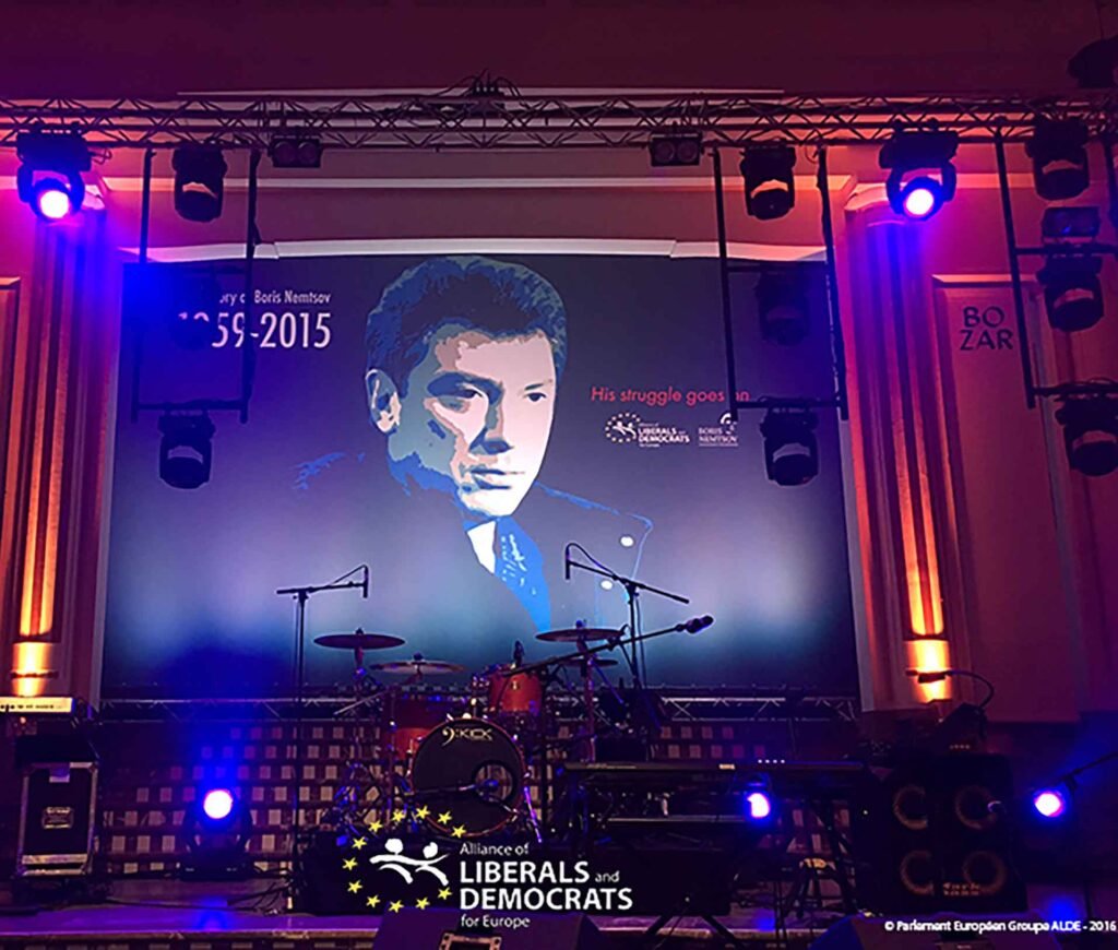 Backdrop en tissu imprimé pour scène de concert, affichant un portrait stylisé d’un homme avec la mention '1959–2015' et 'His struggle goes on', installé dans un lieu de spectacle éclairé par des projecteurs.