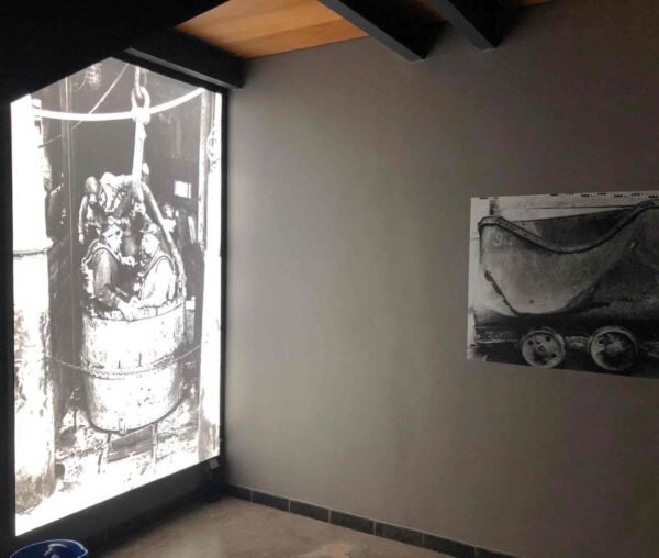 Impression grand format en noir et blanc représentant des mineurs montant dans une cage d'ascenseur, accompagnée d'une image d'un wagonnet de mine sur un mur gris.