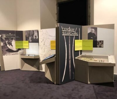 Titre d’exposition intitulé « Cercare / Searching » en lettres découpées en dibond métalliques brossé tridimensionnelles, affiché sur un fond sombre orné d’images de pointes de flèche. À côté, une vitrine contient des artefacts préhistoriques disposés sur une surface claire. Des panneaux jaunes fournissent des informations complémentaires. En arrière-plan, une autre section intitulée « Fotografia / Photography » montre des photos d’archéologues en action.