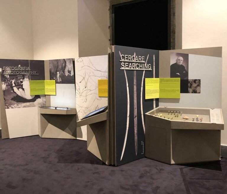 Titre d’exposition intitulé « Cercare / Searching » en lettres découpées en dibond métalliques brossé tridimensionnelles, affiché sur un fond sombre orné d’images de pointes de flèche. À côté, une vitrine contient des artefacts préhistoriques disposés sur une surface claire. Des panneaux jaunes fournissent des informations complémentaires. En arrière-plan, une autre section intitulée « Fotografia / Photography » montre des photos d’archéologues en action.