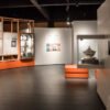 Salle d’exposition muséale avec murs décorés de panneaux informatifs sur fond orange, vitrines d’objets historiques et présentation structurée en modules salle de Musée.