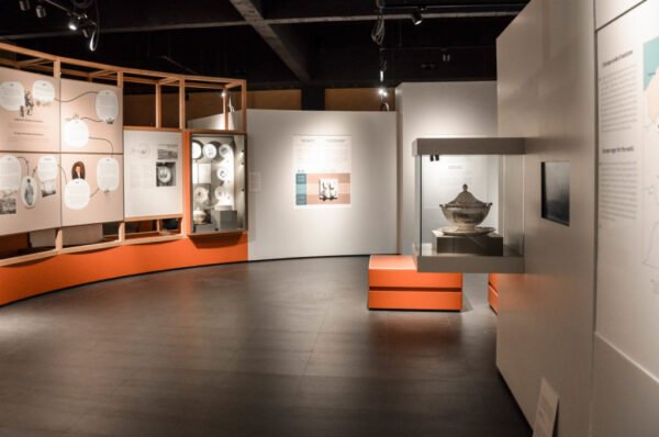 Salle d’exposition muséale avec murs décorés de panneaux informatifs sur fond orange, vitrines d’objets historiques et présentation structurée en modules salle de Musée.