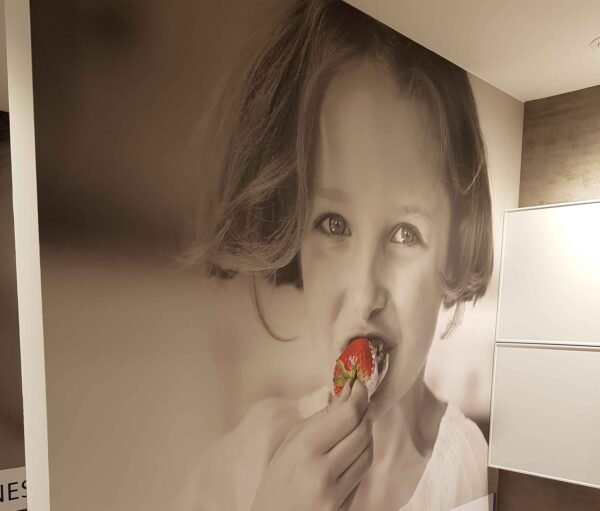 Toile murale en une seule pièce imprimée en grand format, représentant une jeune fille mangeant une fraise, installée dans un espace intérieur moderne pour Batibouw
