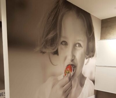 Toile murale sans joint – Photo artistique en une seule pièce Toile murale en une seule pièce imprimée en grand format, représentant une jeune fille mangeant une fraise, installée dans un espace intérieur moderne pour Batibouw
